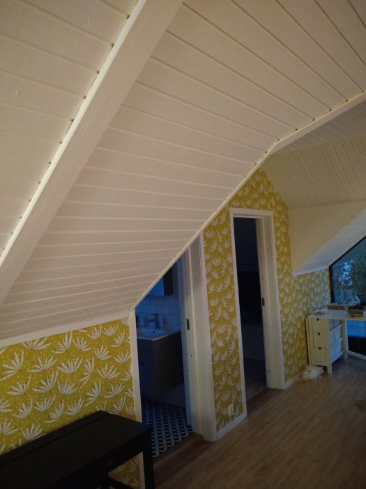 Nyrenoverat vardagsrum med snedtak och ljusa paneler, gulmönstrad tapet och synlig dörröppning till badrummet.