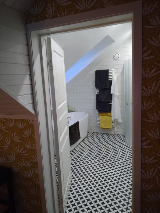 Vy genom en öppen dörr till ett badrum med geometriskt mönstrat golv, vita kakelväggar och ett fristående badkar.