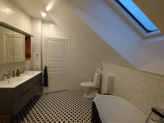 Nyrenoverat badrum med vita kakelväggar, dubbelho, toalett och snedtak med fönster.