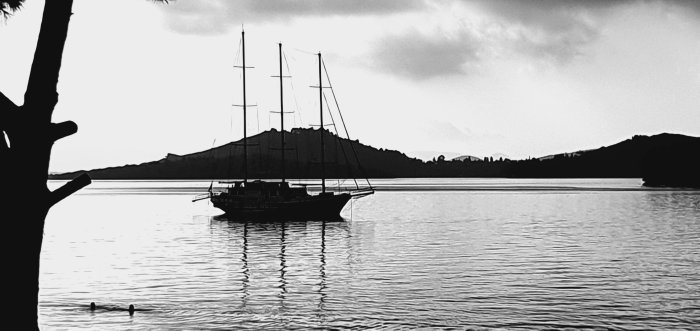 Svartvit bild av en segelbåt på vattnet med berg i bakgrunden och reflektioner.