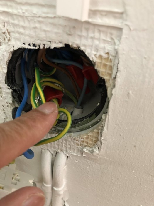 En hand håller en gul/grön jordningskabel nära en öppen eldosa med flera kablar och anslutningar.