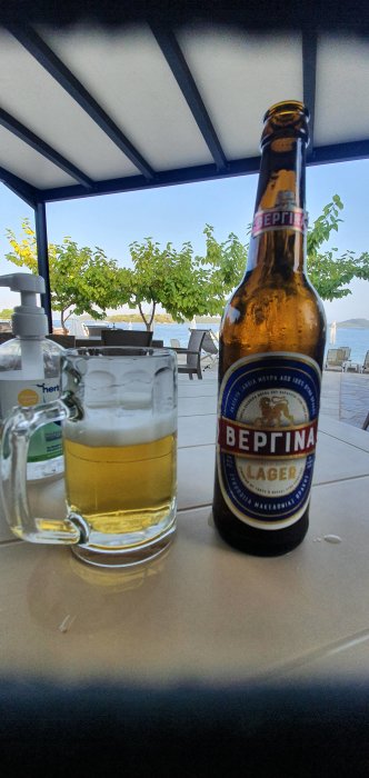 Flaska av Vergina öl bredvid ett glas halvfyllt med öl, utomhusscen i bakgrunden.