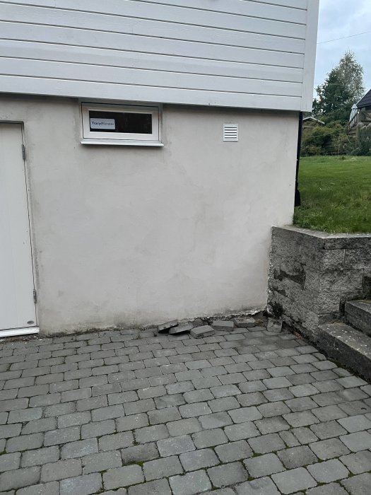 Husvägg med fönster och ventilation, mark med stenläggning och synlig skarv nära husknut med stuprör.
