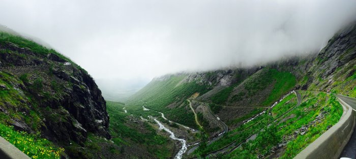 Panoramavy över svingande bergsväg med frodig grönska och lågt molntäcke.