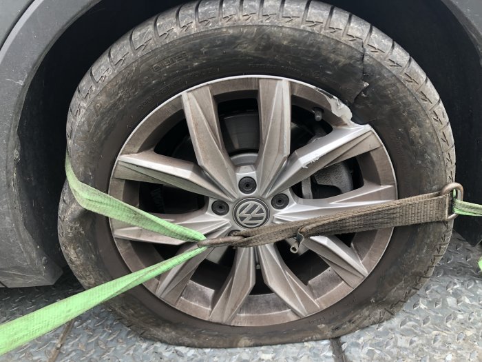 Punkterat bilhjul med slitage och skador, fastspänt med grönt spännband på fordon.
