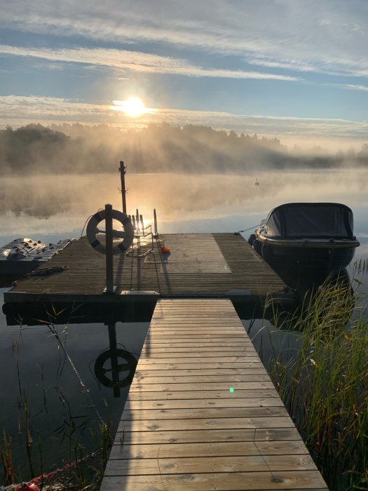 En brygga som leder ut till en dimmig sjö vid soluppgång med en båt kallad "Grodan".