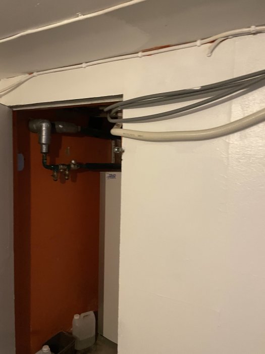 Öppen elcentral med synlig kabeldragning och rörinstallationer vid en vit vägg.
