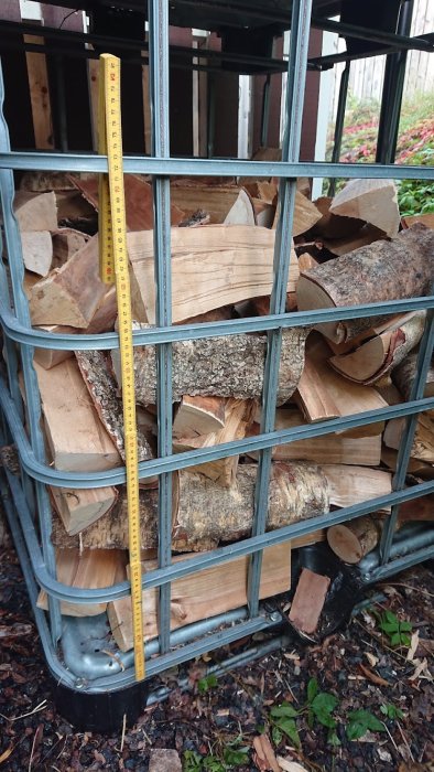 Mätsticka visar höjd på vedstapel i metallbur, ungefär 70 cm hög.