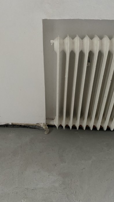 Vattenburen vit radiator i ett rum med grått golv och delvis målad vägg, synliga anslutningsrör i nederkanten.