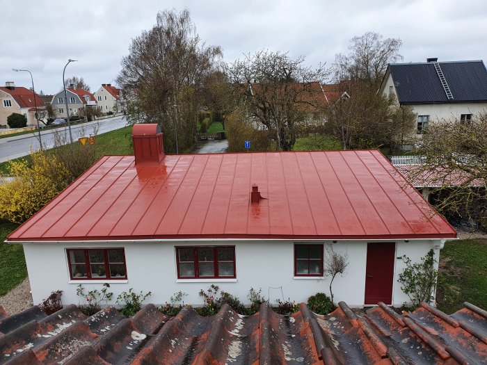 Rött bandfalsat tak på ett vitt hus med tegeltak i förgrunden och trädgård i bakgrunden.