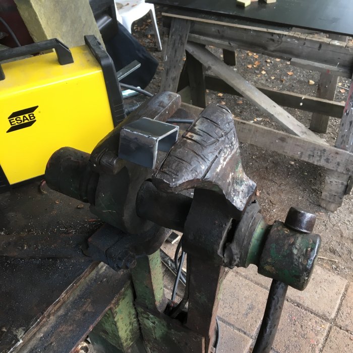 Metallbit fastklämd i en skruvstycke bredvid en gul ESAB svets, arbetsbänk och verktyg i bakgrunden.