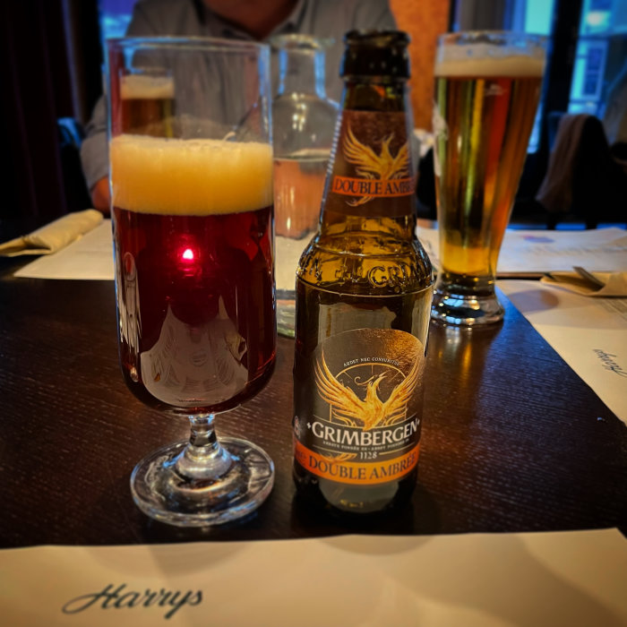 Glas med rödbrun öl och Grimbergen Double Ambré flaska på bord, i bakgrunden otydliga personer.