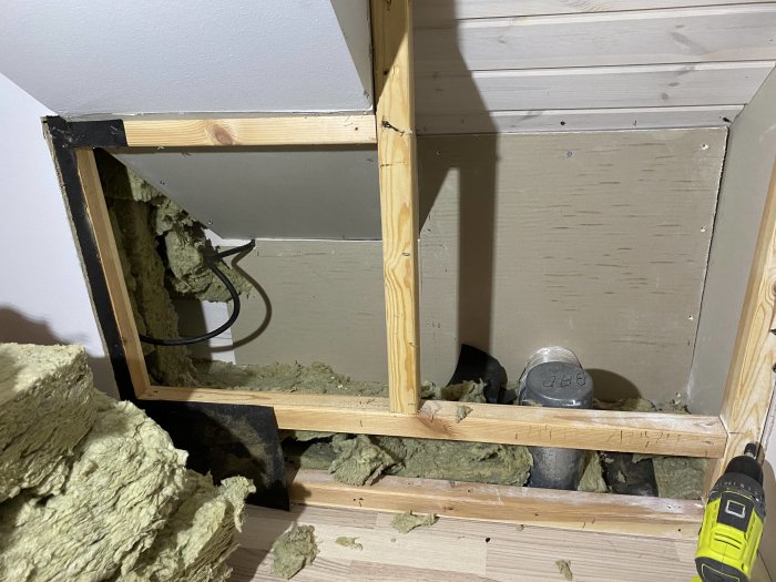 Öppet väggutrymme med isolering och avsaknad av ångspärr på en vind, synlig elledning och spånskiva.