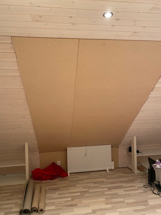 Rum med skevt tak, oskyltad takkupa och golvläggning samt synlig radiator och rullar med väggpapper.