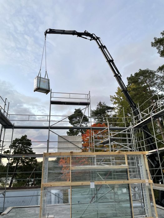 En fasadställning med en lastbilskran som lyfter en byggmodul mot en klarblå himmel.