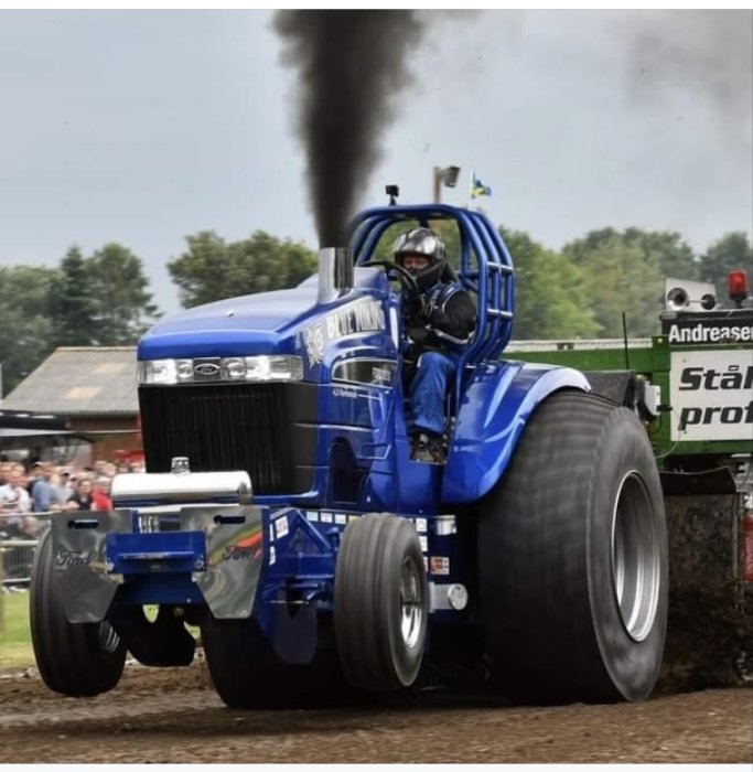 Traktorpulling med blå traktor som släpper ut rök under tävling, publik i bakgrunden.