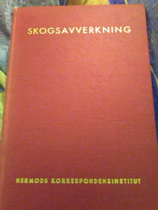 Röd bok med guldtexten "SKOGSAVVERKNING" och "HERMODS KORRESPONDENSINSTITUT" på omslaget.