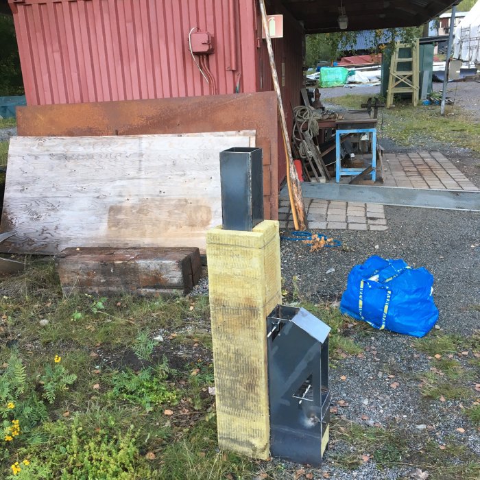 Handgjord asklåda med vinklat galler och metallrör vid en industrilokal, verktyg och material i bakgrunden.