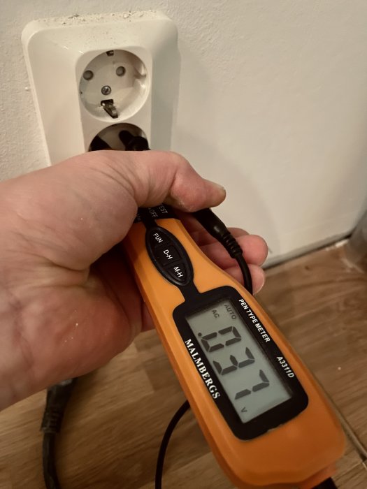 Hand som håller en spänningsprovare mot en vägguttag, visar 231,7 volt.