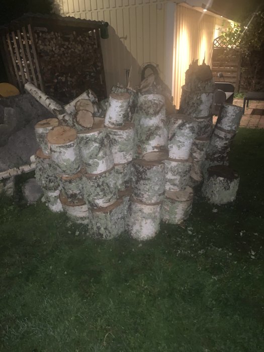 Hög med olagad björkved i trädgård kvällstid, med vedstapel i bakgrunden.