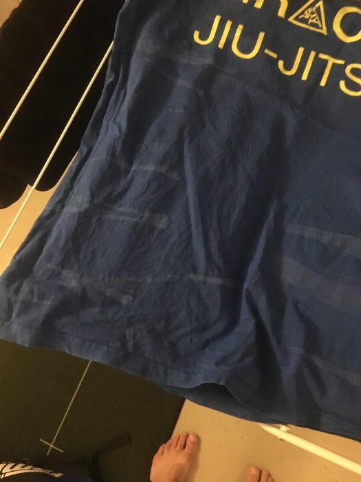Mörkblå t-shirt med text och logotyp ligger på golvet, vita fläckar syns, personens bara fötter i nedre bildkanten.