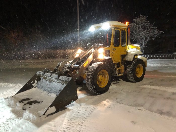 Gul Volvo BM 4200 hjullastare med planeringsskopa som röjer snö på en gårdsplan i snöfall på natten.