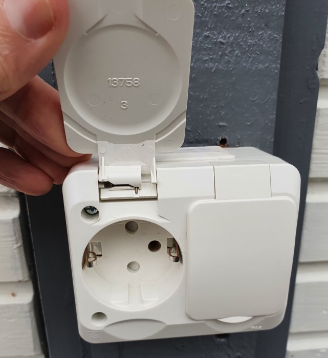 Hand håller ett öppet vitt vägguttag med lock och ospecificerad märkning mot en blå vägg.