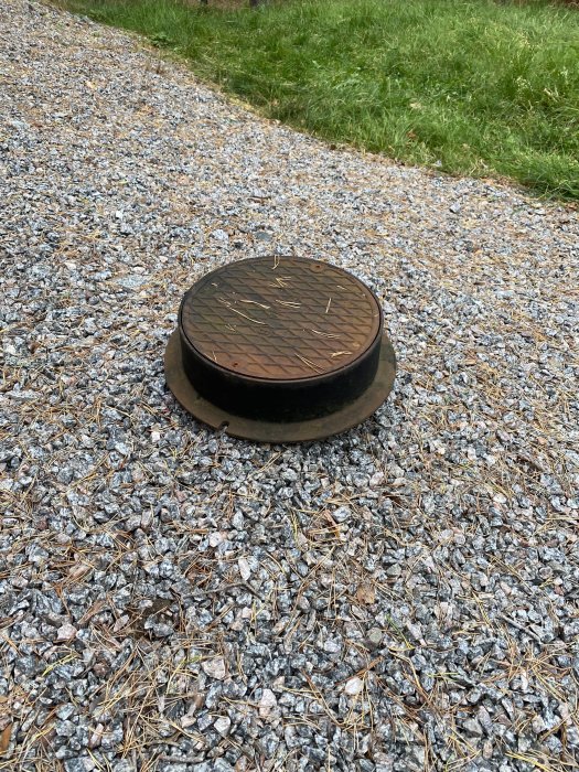 Avloppsledningens inspektionskanal med olåst lock på grusmark.