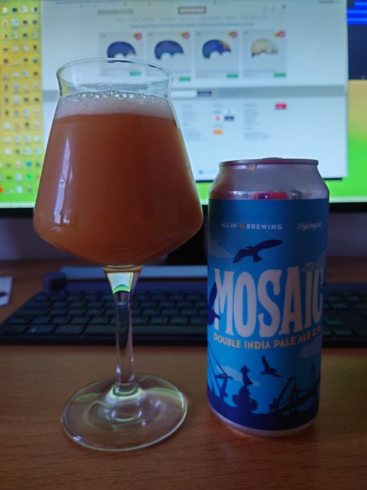 Ett glas med öl bredvid en burk av Mosaic Double India Pale Ale på ett skrivbord framför en datorskärm.