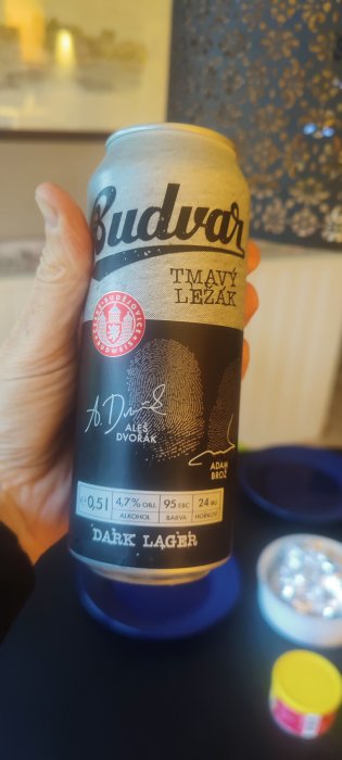 Hand håller en burk Budvar Tmavý Ležák, en mörk lager från Tjeckien, framför en oskarp inredning.