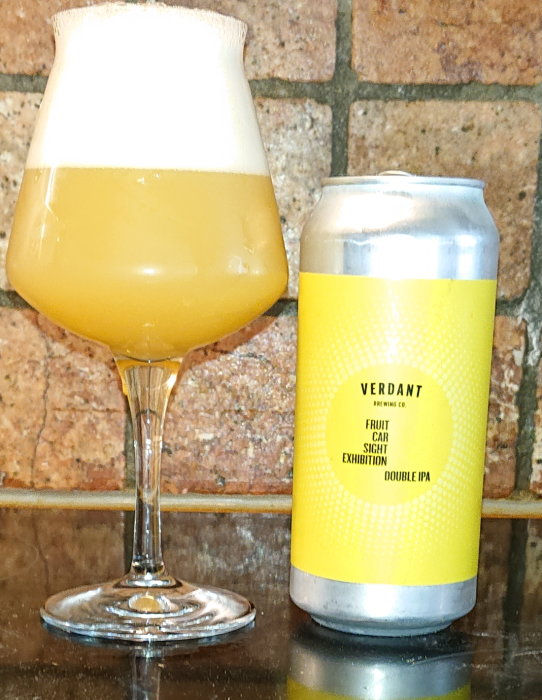 Ölglas fyllt med skummande öl bredvid en burk Double IPA mot tegelvägg.