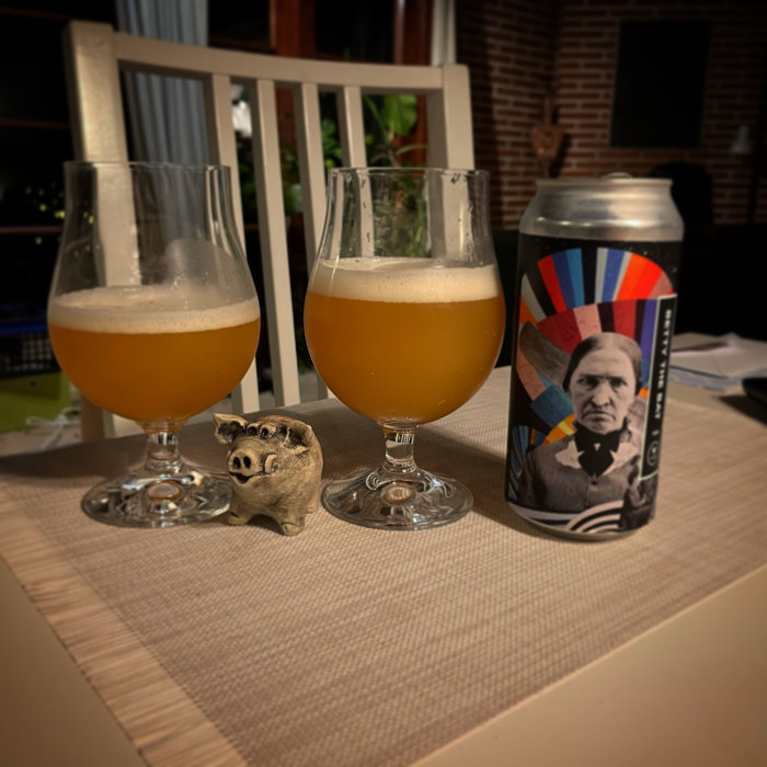 Två glas öl med skum och en ölburk med färgglatt mönster och ansikte, samt en liten grisfigur på bord.