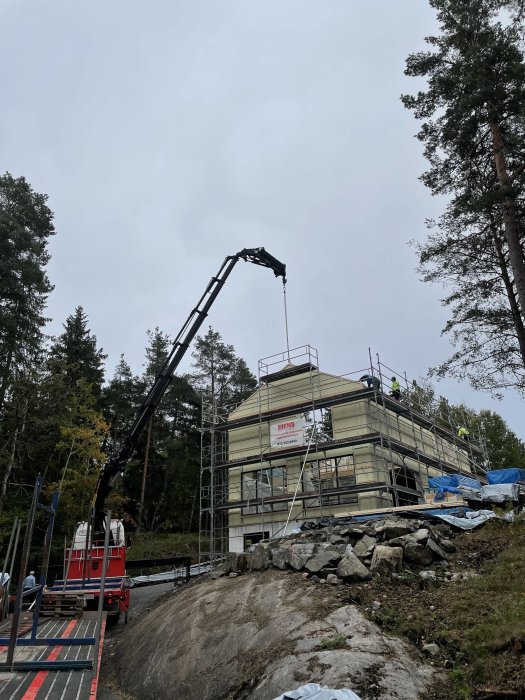 Hus under konstruktion med ställningar, råspont på taket och en kranbil, omgiven av skog och klippor.