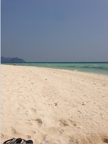 Strandvy med turkost hav och få människor i bakgrunden, fotavtryck i sanden, flip-flops i förgrunden.