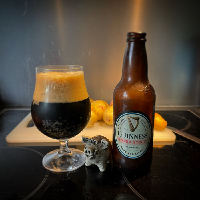 Guinness Extra Stout ölflaska bredvid ett glas mörk öl och en småfigur av en gris på en köksbänk.