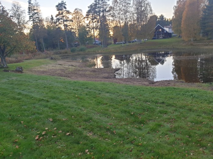 Nygrävda kanter vid en damm med gräsmatta i förgrunden och hus i bakgrunden under hösttid.