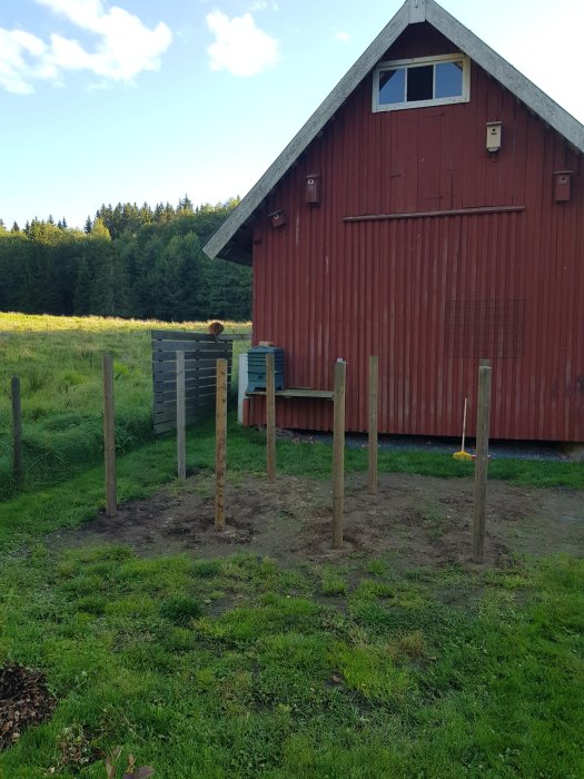 Uppställda trästolpar som markerar starten på en ny trädgårdskompost framför ett rött lantligt uthus.