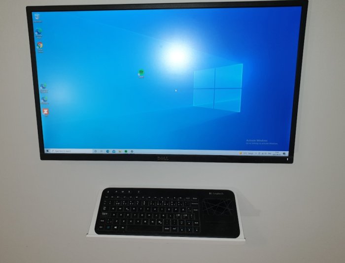 Datorskärm på vägg med Windows-skärmbild ovanför en hylla med trådlöst tangentbord och mus.