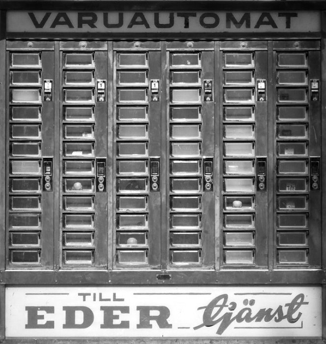 Svartvit bild av en gammal varuautomat med många små luckor, använd för köp av varor utanför arbetstid.