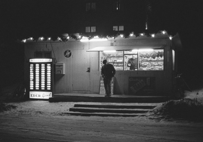 Svartvit bild av en kiosk öppen på kvällen med en kund och en automat vid sidan om.