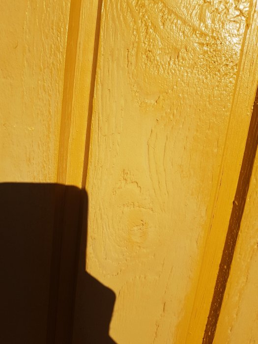 Närbild av gulockra linoljefärg på husfasadens träpanel med synlig träådring och skugga.