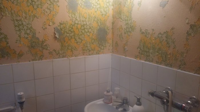 Äldre badrum med beige tapet med gul blomstermotiv ovanför vita kakelplattor, en tvättställsblandare och T-ventiler.