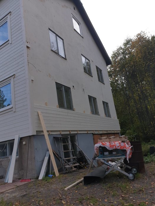 Delvis renoverat hus med ny träpanel under fönsterna och byggmaterial spritt omkring på marken.