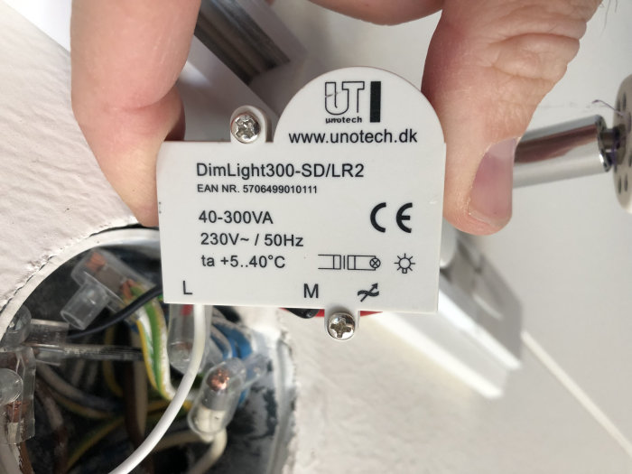 En person håller en DimLight300-SD/LR2 dosdimmer mot en vägg med synliga elektriska trådar och anslutningar i bakgrunden.