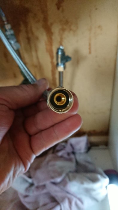 Hand håller en sprucken slanganslutning för kran med balofix i bakgrunden.