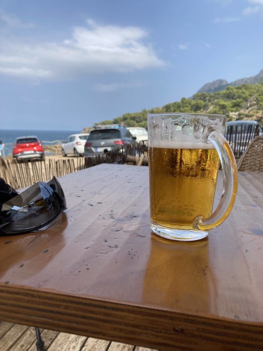 Ölglas på träbord med solglasögon och havsutsikt i bakgrunden.