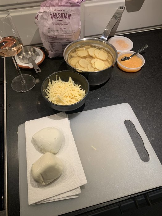 Ingredienser för Bianco med potatis och löjrom förbereds på köksbänk, inklusive två mozzarellaostar, rivet ost, och ett glas vin.