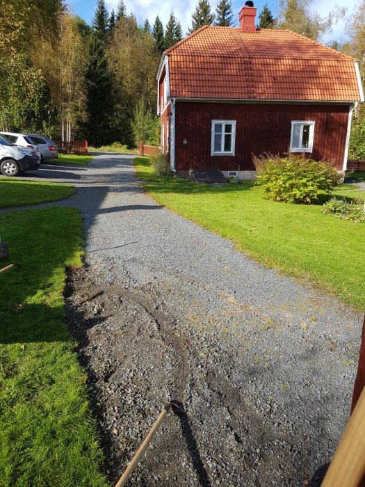 Nyanlagd kant längs infarten med grus och rådjurmotiv, traditionellt rött hus i bakgrunden.