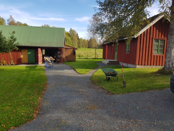 Grusad infart med kantning fram till parkering och gång, röda stugor och skottkärra i trädgården.