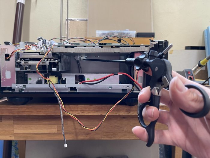 Öppnad hembioprojektor med synliga interna komponenter och kablar bredvid en hand som håller en laparoskopitång.